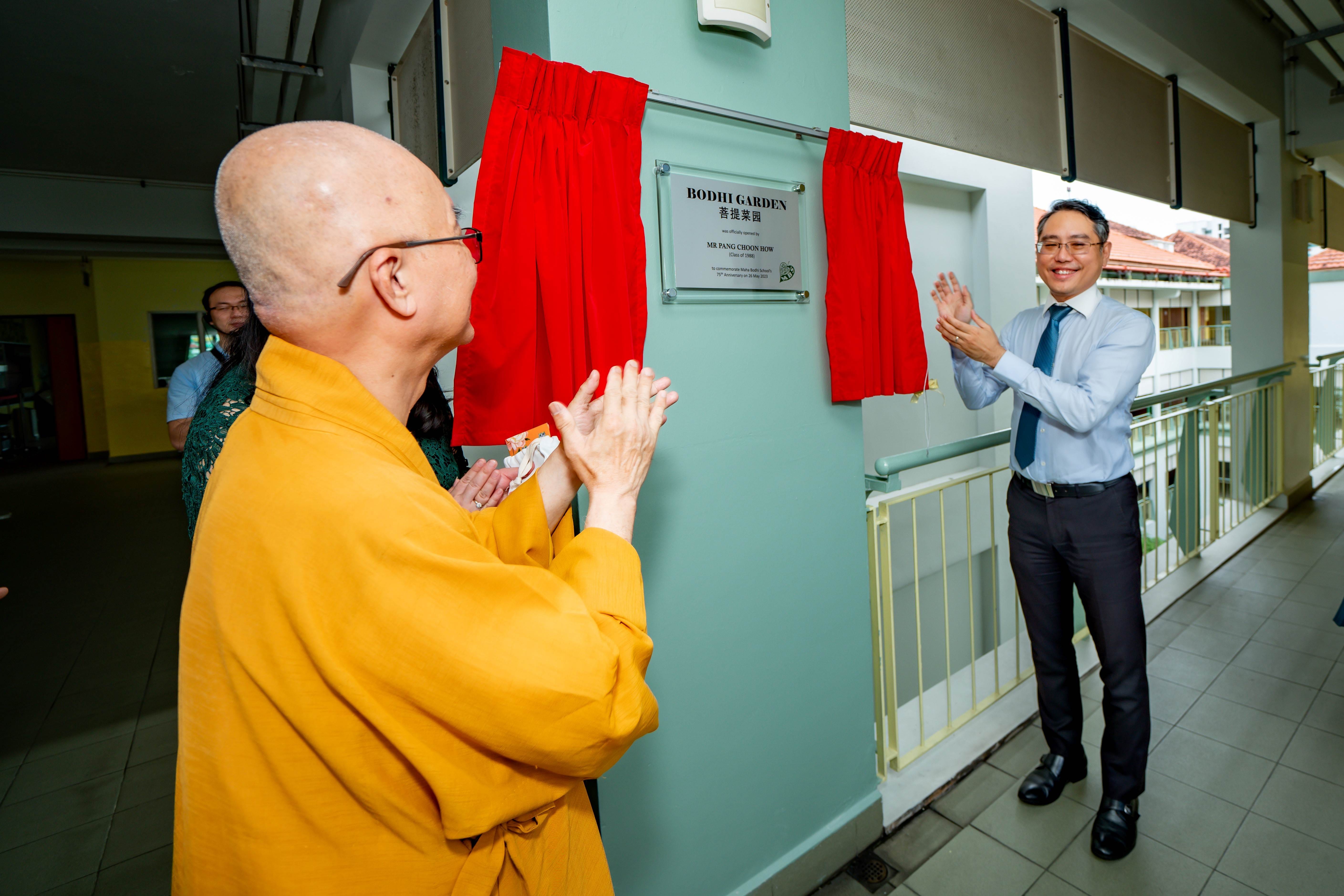A monk in yellow robes and a suited man applaud beside a unveiled Bodhi Garden plaque draped with red curtains.